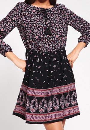 Schwarzes Blumenkleid mit gerafftem Oberteil, langen Ärmeln und Quasten-Detail. Der Rock hat einen gemusterten Saum in verschiedenen Rosa- und Lilatönen.