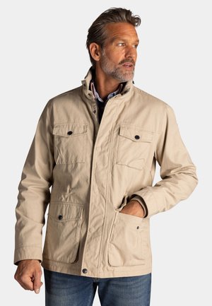 Man draagt een beige utiliteitsjack met vier zakken aan de voorkant, staat met één hand in de zak en kijkt naar rechts.