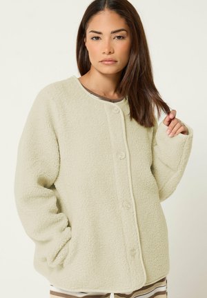 Femme aux cheveux bruns et raides porte une veste polaire crème oversize avec des boutons, posant avec une main dans la poche et l'autre tenant ses cheveux.
