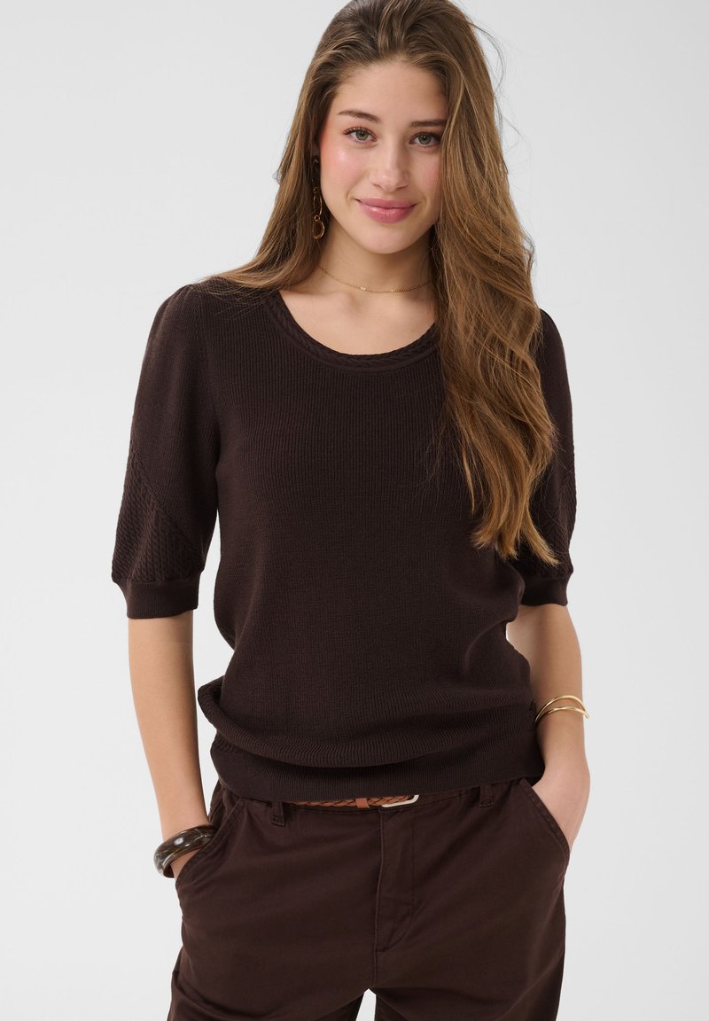 Jeune femme aux longs cheveux châtain clair portant un pull à manches courtes marron foncé et un pantalon assorti, souriante, les mains dans les poches, sur fond uni.