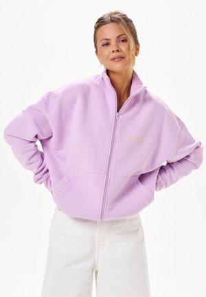 Femme aux cheveux attachés en chignon lâche, portant une veste polaire zippée violet clair et un pantalon blanc, debout, les mains sur les hanches, sur fond blanc.