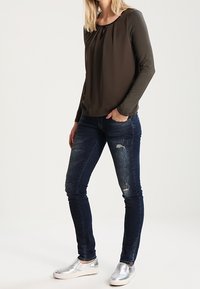 Olivgrön långärmad topp med veckad halsringning, tillsammans med mörkblå slim-fit jeans med slitna detaljer och silverfärgade slip-on skor.