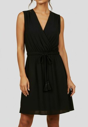 Mouwloze zwarte jurk met V-hals overslagtop, elastische taille en decoratieve kwaststrik, gedragen door een vrouw tegen een effen achtergrond.