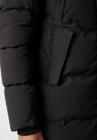 Must must jacket, mille õmblusega tekstuur, pikad varrukad ja rinnatasku lukuga. Silmapaistev kaubamärgi logo detail on nähtav.