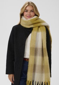 Sciarpa a quadri gialla e bianca con bordi frangiati, realizzata in tessuto morbido, drappeggiata intorno al collo, abbinata a un cappotto nero con texture.