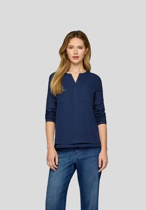 Navy blue top with long sleeves, v-neck, and two front pockets. Smooth fabric with a slight sheen, relaxed fit, paired with blue jeans.