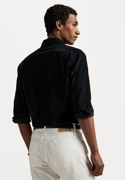 Black corduroy shirt with a relaxed fit, rolled sleeves, and a classic collar, paired with light-colored denim jeans.