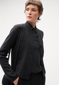 Chaqueta de mezcla de lana gris con un cuello alto, botones frontales y bolsillos laterales. Llevada sobre una parte superior a juego, combinada con pantalones oscuros de canalé.