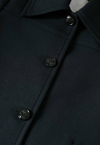 Veste en textile noir avec un col structuré, dotée de cinq boutons noirs brillants et d'un tissu texturé. Coutures détaillées visibles.