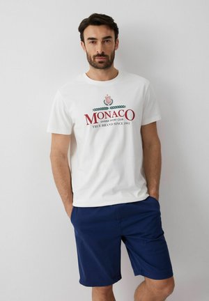 Mężczyzna ubrany w białą koszulkę z napisem „MONACO” i granatowe szorty, stoi z rękami w kieszeniach na jednolitym jasnym tle.