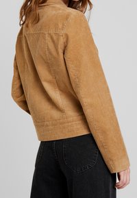 Jasnobrązowa kurtka z korduroju o żebrowanej fakturze, prostej sylwetce i szwach z tyłu. Posiada kołnierz oraz wyraźnie zarysowane ramiona, a także dopasowany pas.