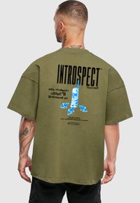 Tricou larg de culoare verde măsliniu, cu un design grafic albastru pe spate, text imprimat și o croială unică, casual.