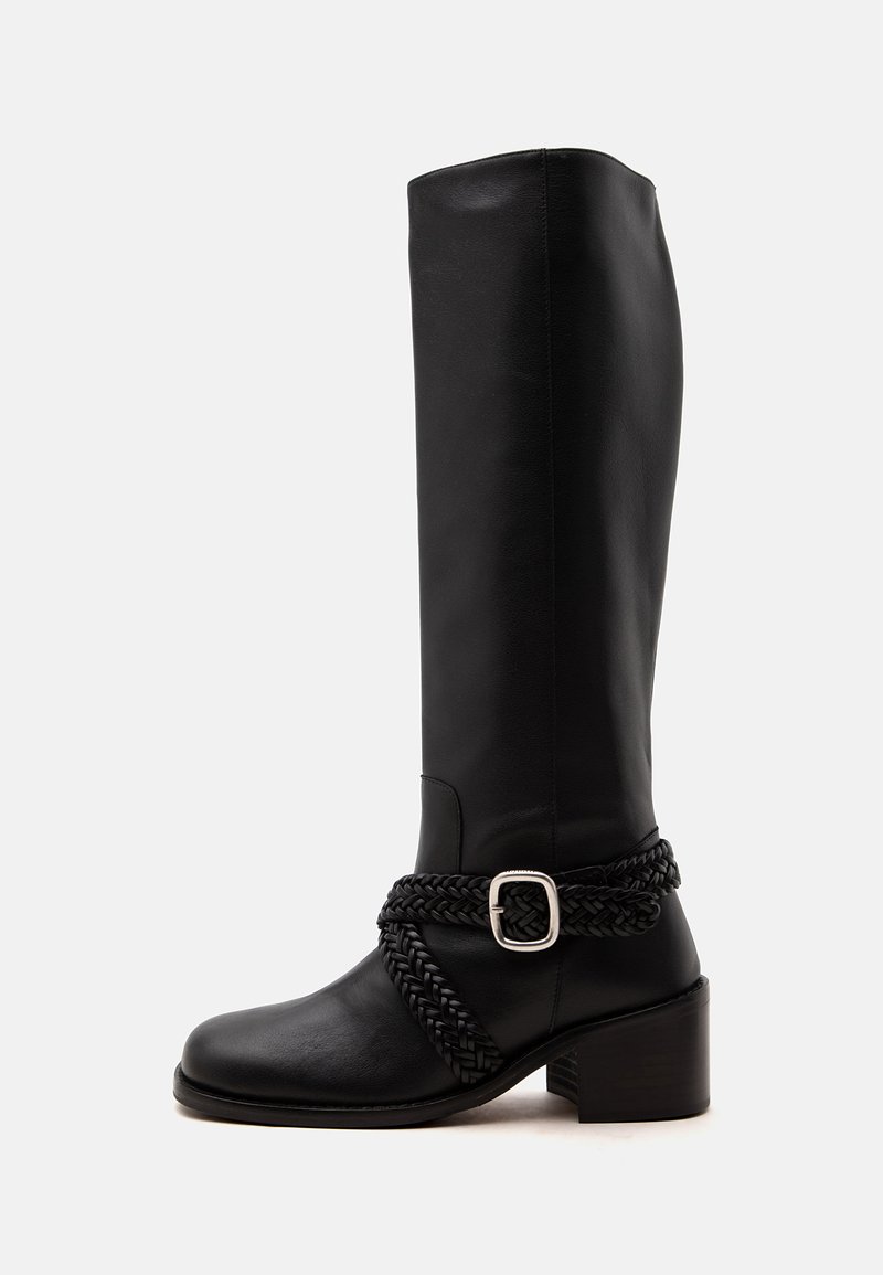 Botte noire en cuir montant au genou avec talon bloc et sangle tressée avec une boucle argentée autour de la cheville.
