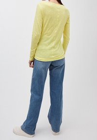 Eine Person, die mit sichtbarem Rücken steht und ein hellgelbes Langarmoberteil, blaue weit geschnittene Jeans und weiße Turnschuhe trägt, vor einem einfarbigen Hintergrund.