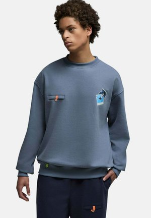 Jeune personne portant un sweat-shirt bleu avec deux impressions photo de skateboard sur la poitrine et un pantalon bleu assorti avec des boutons orange.