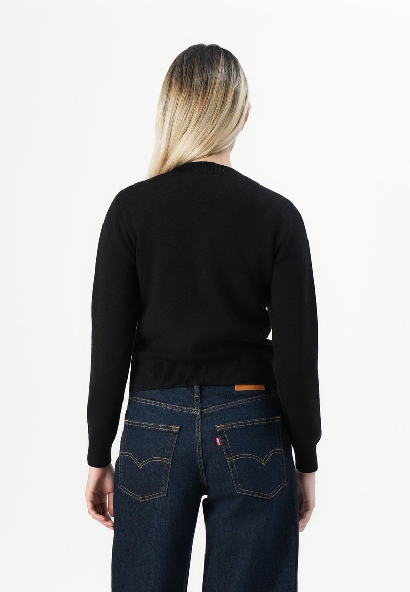 Femme aux longs cheveux blonds portant un haut noir à manches longues et un jean bleu foncé, debout de dos contre un fond blanc.