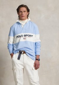 Gaiši zils garām piedurknēm polo krekls ar baltu svītru, uz kura ir uzraksts "POLO SPORT" jūras zilā krāsā. Apvienots ar baltām cargo biksēm un daudzkrāsainu jostu.