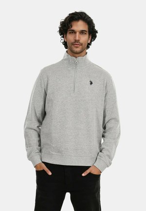 Hellgrauer Zip-Hoodie aus weichem Material, mit hohem Kragen, gerippten Bündchen und einem kleinen schwarzen Logo auf der linken Brust.