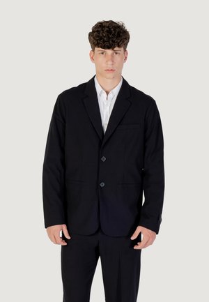 Giovane uomo con capelli ricci, indossa una giacca nera e una camicia bianca, in piedi davanti a uno sfondo chiaro e semplice.