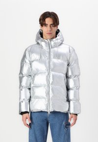 Giacca puffer argento con una finitura lucida e riflettente, cappuccio e chiusura frontale con zip. Presenta un design trapuntato con cuciture e texture visibili.