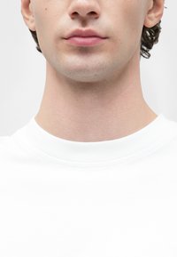 Biele tričko s okrúhlym výstrihom a jemnou textúrou. Výstrih je lemovaný rebrovaným lemom. Pod tričkom je viditeľný krk v odtieni pokožky.