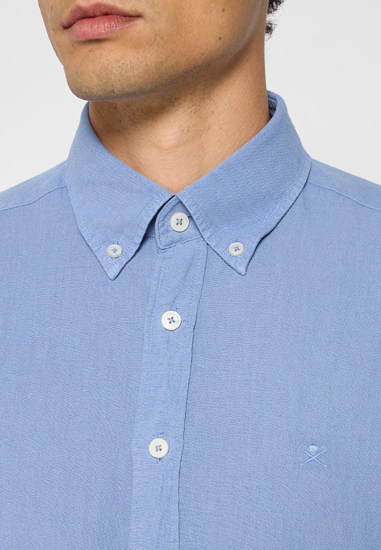Camisa de botones azul claro con un cuello estructurado, pequeños botones blancos y un sutil bordado del logo en el pecho izquierdo. Tejido texturizado.