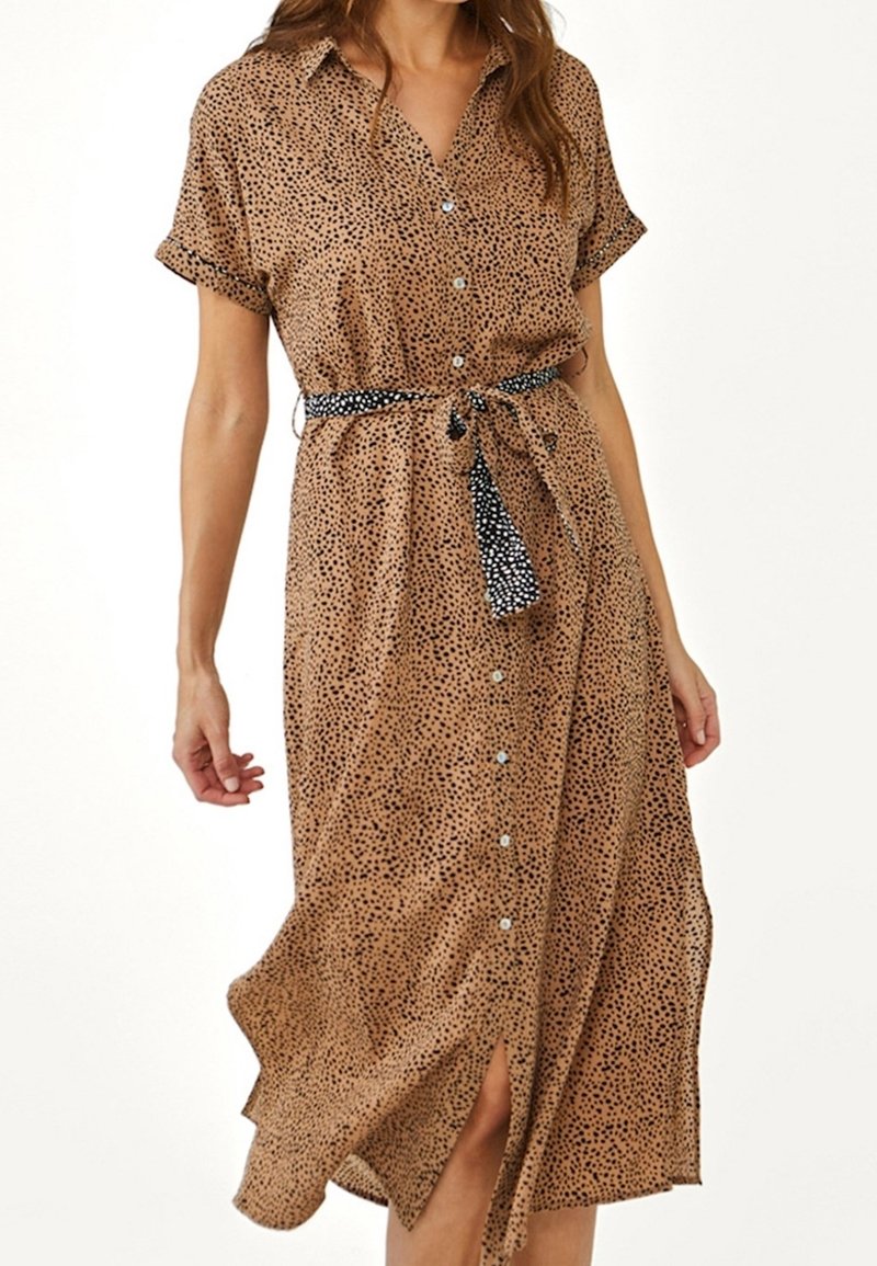 Femme portant une robe marron clair à manches courtes boutonnée avec des taches noires et une ceinture contrastante noire et blanche à pois nouée.