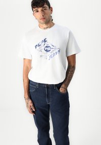 Tatuiruotėmis išmargintas vyras, vilkintis baltą Pepe Jeans marškinėlį ir tamsiai mėlynas džinsus, stovintis prieš šviesiai baltą foną.