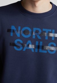 Námořnická modrá mikina s žebrovaným kulatým výstřihem a modrým textem "NORTH SAILS" částečně překrytým dvěma reflexními stříbrnými a černými horizontálními pruhy.