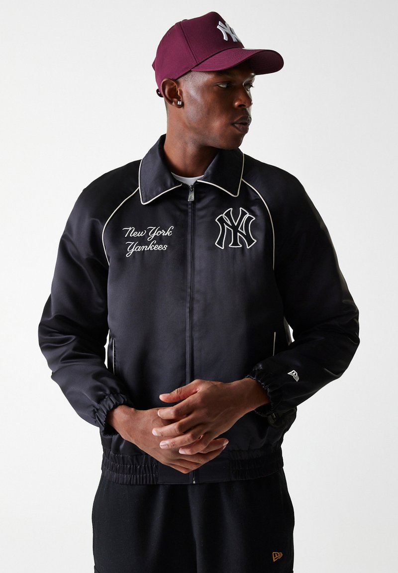 Juodas nailono švarkas su balta siūle, tekstu "New York Yankees" ir siuvinėjimu; turi užtrauktuką priekyje ir elastines rankoves. Bordo spalvos kepurė.