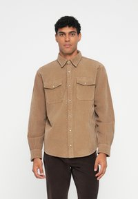 Bruine ribfluweelshirt met een button-down kraag, twee voorzakken en een subtiele verticale ribstructuur. Lange mouwen met knoopmanchetten.