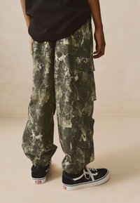 Green and black cargo pants with a splatter pattern, featuring multiple pockets, elastic cuffs, and a relaxed fit, paired with black sneakers.