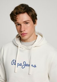 Felpa bianca con logo ricamato "Pepe Jeans" blu. Il tessuto sembra morbido, con tasca a marsupio e coulisse regolabile.