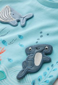 Svetlomodrá košeľa s vyšívanými grafikami veľryby a rýb, s textúrovanými nášivkami usmievajúceho sa vodného tvora a detailmi koralu.