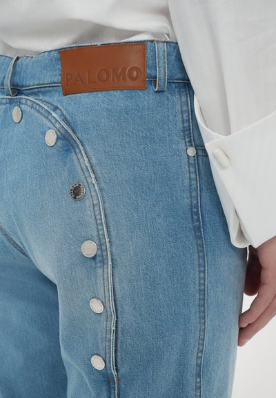 Jeans din denim de culoare albastră deschisă, prezentând nasturi metalici pe partea laterală și o etichetă din piele maro cu inscripția "PALOMO" pe talie.