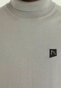 Pull à col roulé gris en tissu doux avec texture côtelée, coupe slim, et petit patch logo noir sur la poitrine gauche.