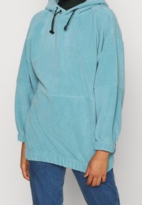 Persona che indossa un hoodie in pile blu chiaro con lacci e polsini elastici, abbinato a jeans in denim blu su uno sfondo semplice.