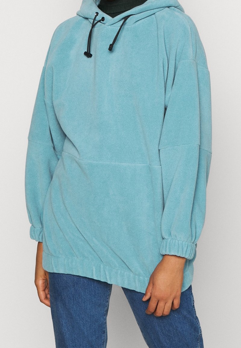 Persona che indossa un hoodie in pile blu chiaro con lacci e polsini elastici, abbinato a jeans in denim blu su uno sfondo semplice.