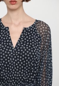 Blouse bleu marine avec de petits motifs floraux blancs, décolleté en V et manches longues transparentes, dotée d'une ceinture élastique pour un ajustement parfait.