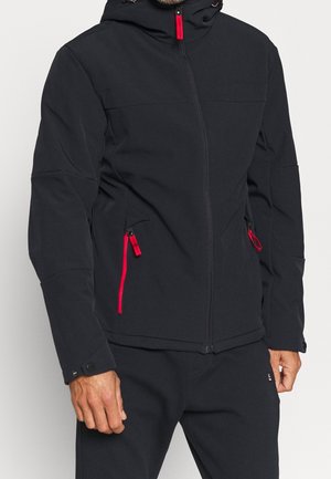 Mann trägt eine schwarze Jacke mit Reißverschluss und roten Zippern sowie schwarze Hose, steht vor hellem schlichten Hintergrund.