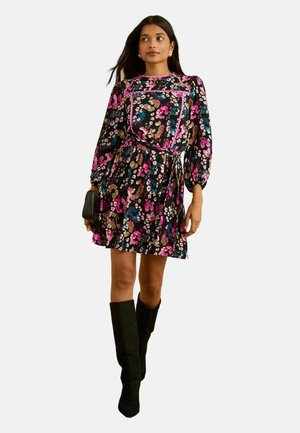 Frau trägt ein knielanges florales Kleid mit rosa, blauen und braunen Mustern, kombiniert mit schwarzen kniehohen Stiefeln und hält eine kleine schwarze Clutch.