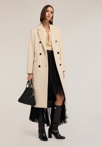 Cappotto in misto lana beige con bottoni neri, indossato sopra una blusa gialla e una gonna nera a strati. Indossato con stivali neri a ginocchio in tessuto texture e una borsa.