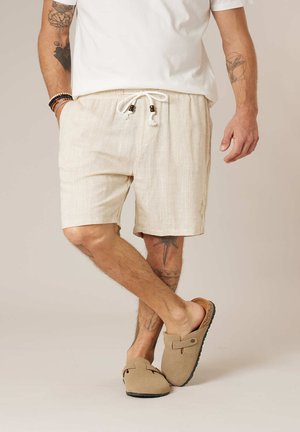 Homme portant un short beige à cordon, un t-shirt blanc, des chaussures plates beiges et des bracelets, debout jambes croisées devant un fond neutre.