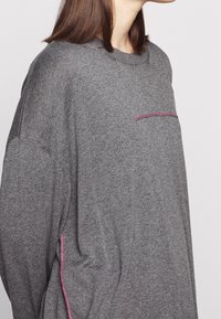 Gray oversized sweater made from soft material, featuring a pink accent stitch on the chest pocket and side seam. Crew neckline, loose fit.