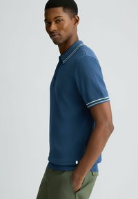 Marineblaues Poloshirt mit weicher Textur, zwei Knöpfen und einem gerippten Kragen mit weißen Streifen. Der Schnitt ist tailliert und hat kurze Ärmel.