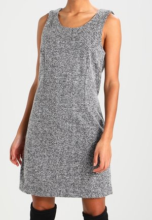 Szara sukienka bez rękawów z wzorem jodełki, sięgająca do kolan, noszona z czarnymi wysokimi kozakami na stojącej osobie.