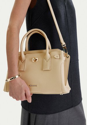 Béžová texturovaná kožená kabelka se zlatými kovovými doplňky a logem, držená osobou v černém topu bez rukávů a se zlatým náramkem.