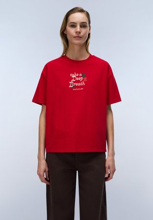 Kvinde iført en rød T-shirt med teksten ""take a Deep Breath"" og en lille grafik, parret med mørkebrune bukser, stående foran en ensfarvet baggrund.