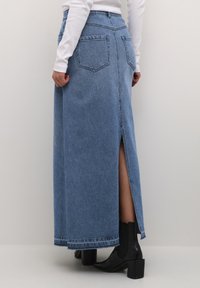 Denim maxi skirt, light blue, features a high slit on the left side, traditional five-pocket design, and frayed hem. Worn with black ankle boots.