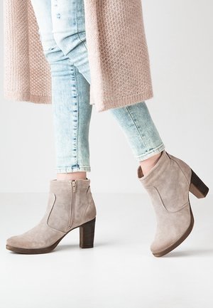 Bottes de cheville en daim beige avec une fermeture éclair sur le côté, un design incurvé et un talon empilé marron, associées à un jean bleu clair déchiré.